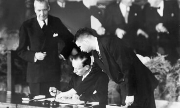 Henrik Kauffmann (1888-1963) er en af de mest betydningsfulde og gådefulde skikkelser i dansk diplomati. På dette billede fra 1949 ses han som Danmarks ambassadør i USA stående i baggrunden, mens den daværende danske udenrigsminister Gustav Rasmussen underskriver Atlanterhavspagten assisteret af John W. Foley (TH), en amerikansk embedsmand.